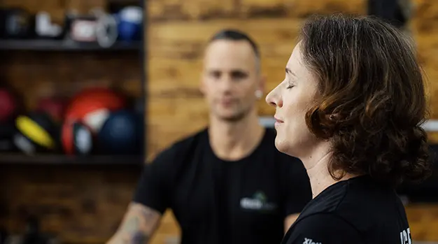 Eine Frau sitzt mit geschlossenen Augen in einem Fitnessstudio in Augsburg, während Marco Neumann im Hintergrund eine Atemtechnik anleitet.