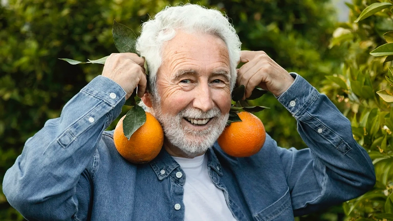 Ein älterer Mann zeigt mit einem Lächeln zwei Orangen als Sinnbild für gesund altern durch vitaminreiche Ernährung und Lebensfreude